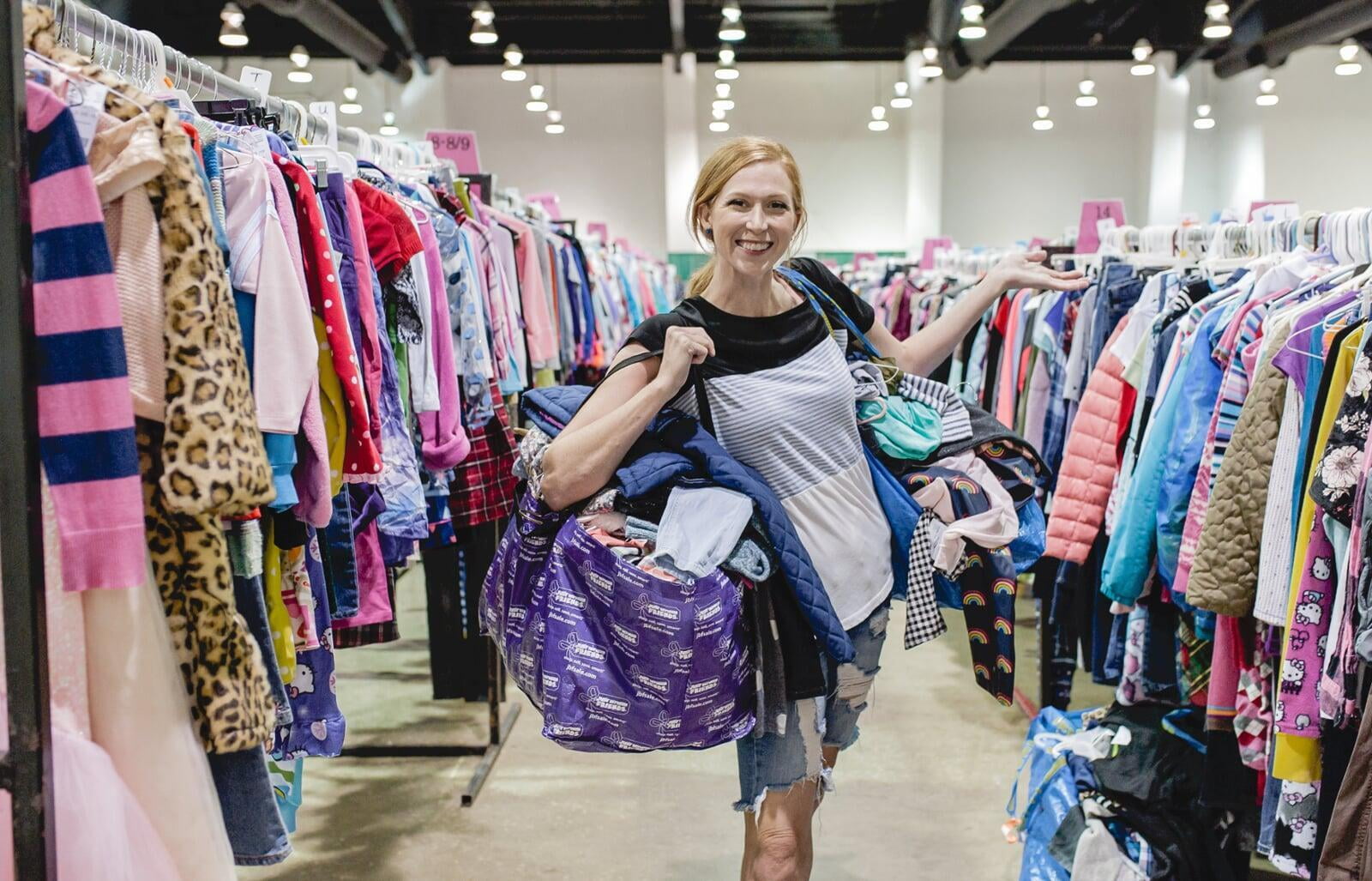 A mom with a large JBF shopping bags on her shoulders stand between clothing racks at JBF!