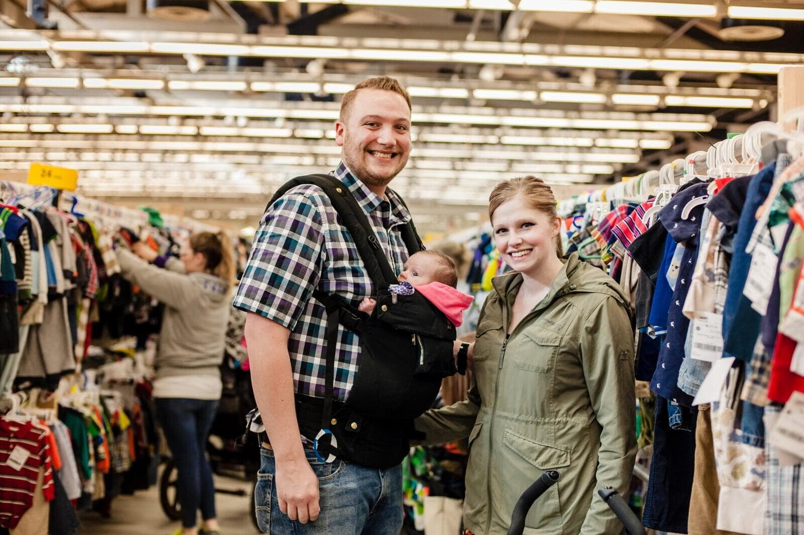 mother and father shopping in an aisle of clothes.  Father is baby wearing an infant